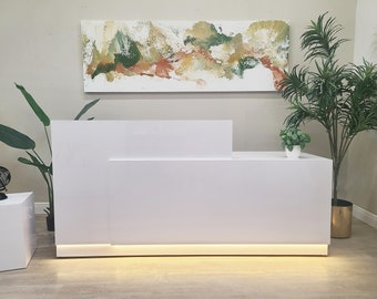 May include: White modern reception desk with a white countertop and a white base. The desk has a built-in light strip along the bottom edge. There is a painting on the wall behind the desk, and a potted plant on the right side of the desk.
