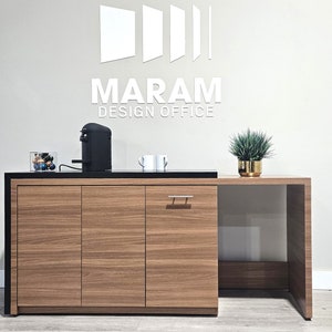 May include: A black and wood office coffee station with a black coffee maker, a glass jar of coffee pods, two white mugs, and a small potted plant. The station has a black countertop and a wood cabinet with three doors. The wall behind the station has a white logo that says "MARAM DESIGN OFFICE".