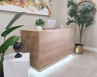 May include: A light wood reception desk with a gold trim, a laptop computer, and a potted plant on top. The desk is illuminated by a strip of LED lights underneath.