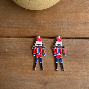 May include: A pair of red, white and blue nutcracker earrings. The nutcrackers are smiling and have black moustaches. The earrings are made of metal and have a shiny finish.