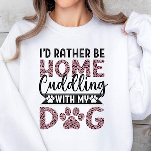 May include: White sweatshirt with the text "I'd rather be home cuddling with my dog" in black lettering. The word "home" and the word "dog" are printed in a pink leopard print.