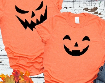 May include: Two orange t-shirts with black jack-o'-lantern faces. One shirt has an angry face, and the other has a smiling face. The shirts are on a white wooden surface with autumn leaves.
