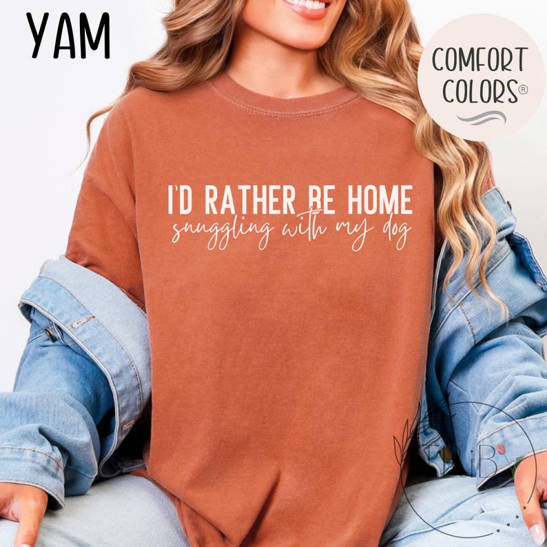 May include: Rust-colored t-shirt with the text "I'D RATHER BE HOME snuggling with my dog" in white script. The shirt is part of the Comfort Colors collection and is paired with a denim jacket.