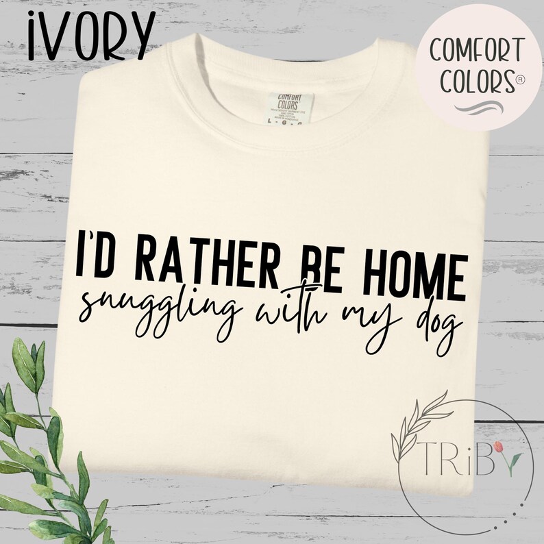 May include: Ivory-colored t-shirt with the text "I'D RATHER BE HOME snuggling with my dog" in black. The shirt is a Comfort Colors brand. The design includes a small leaf graphic and the word "TRIBY".