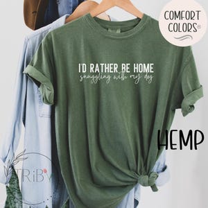May include: A green t-shirt with the text "I'D RATHER BE HOME snuggling with my dog" in white. The shirt is hanging on a wooden hanger with other clothing items in the background. The shirt has rolled-up sleeves.