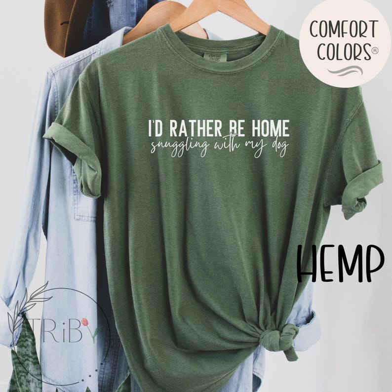 May include: A green t-shirt with the text "I'D RATHER BE HOME snuggling with my dog" in white. The shirt is hanging on a wooden hanger with other clothing items in the background. The shirt has rolled-up sleeves.
