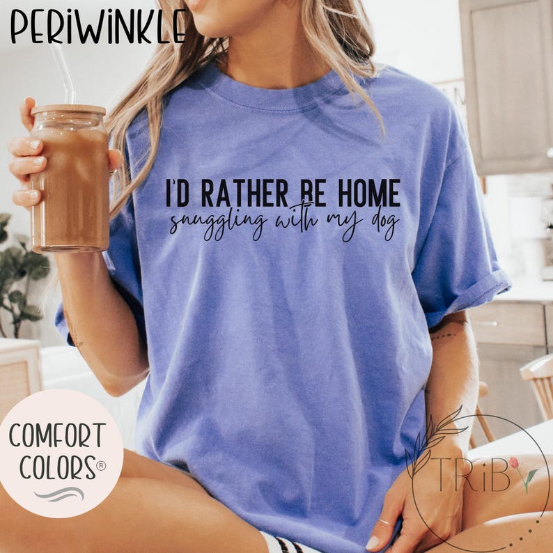 May include: Periwinkle Comfort Colors t-shirt in a periwinkle blue hue. The shirt features the text "I'd rather be home snuggling with my dog" in a black script. The shirt is a relaxed fit.