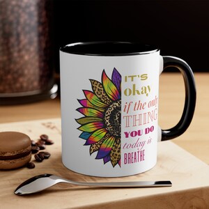 May include: White coffee mug with a black rim and handle. The mug features a colorful sunflower with a leopard print design and the text "It's okay if the only thing you do today is breathe."
