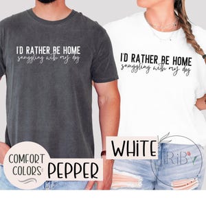 May include: Two t-shirts, one pepper and one white, with the text "I'D RATHER BE HOME snuggling with my dog" in a script font. The pepper shirt has "COMFORT COLORS" and "PEPPER" printed on it. The white shirt has "WHITE" printed on it.