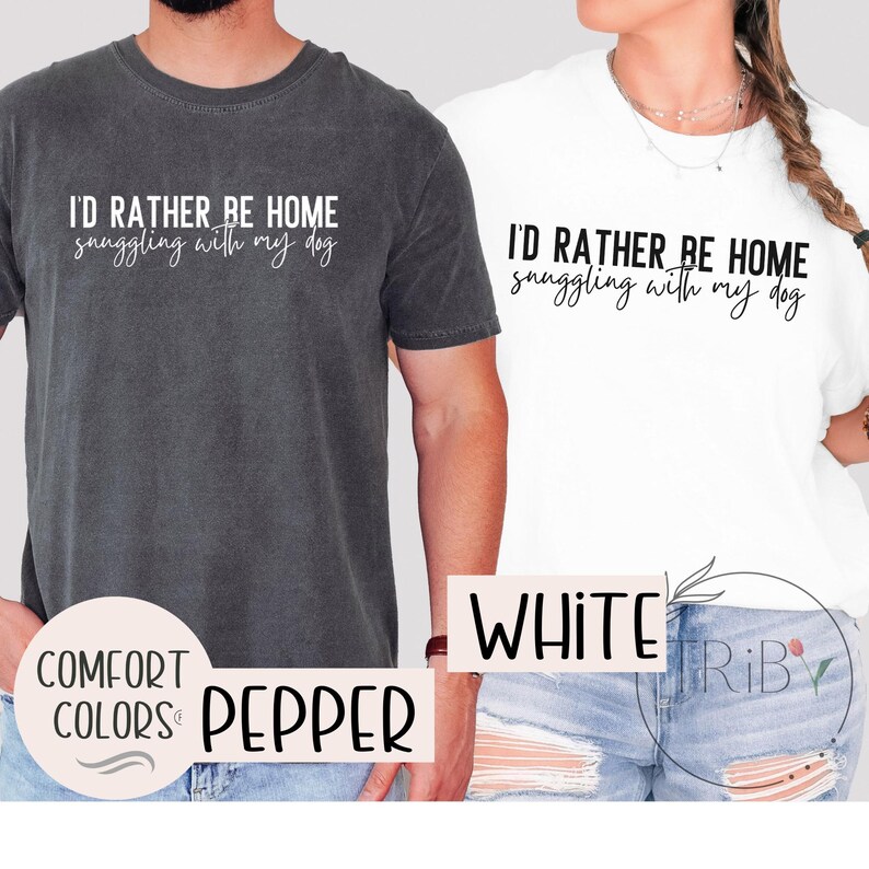 May include: Two t-shirts, one pepper and one white, with the text "I'D RATHER BE HOME snuggling with my dog" in a script font. The pepper shirt has "COMFORT COLORS" and "PEPPER" printed on it. The white shirt has "WHITE" printed on it.