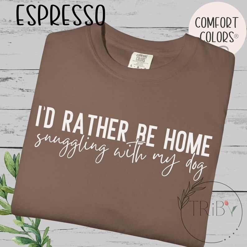 May include: Espresso-colored t-shirt with the text "I'd rather be home snuggling with my dog" in white script. The shirt has a crew neck and a Comfort Colors tag. The shirt is on a light wood-grain background.