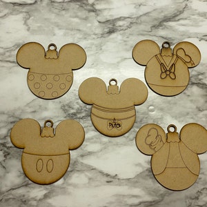 Op de afbeelding: Vijf onafgewerkte houten Mickey Mouse-hoofdornamenten. Eén heeft stippen, een andere heeft de naam Pluto, en de anderen tonen Mickey, Donald Duck en een personage met een hoed. Elk heeft een kleine lus om op te hangen.