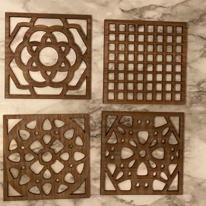 May include: Four wooden coasters with intricate laser-cut designs. The coasters feature geometric patterns, including circles, squares, and triangles. The coasters are made of a dark brown wood and have a natural finish.