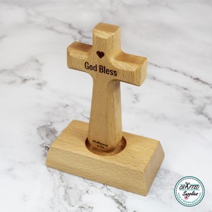 May include: A wooden cross with the words "God Bless" engraved on it. The cross is standing on a wooden base and has a small heart cutout in the center. The base has the words "Philippians 4:13" engraved on it.