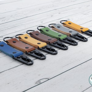 May include: Seven leather keyrings with metal key rings and clips in various colours, including blue, brown, yellow, green, and grey. The keyrings are arranged in a row on a white wooden surface.