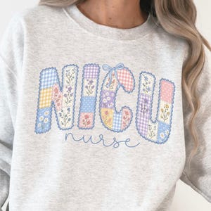 May include: An ash grey crewneck sweatshirt featuring the text NICU in a patchwork style with blue, pink, and yellow floral and gingham patterns. Below the large letters, the word nurse is written in a simple blue cursive script font.