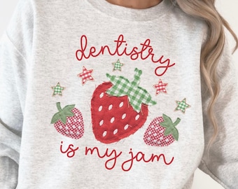 Puede incluir: Una sudadera gris ceniza de cuello redondo con un gráfico de tres fresas con estampado de cuadros rojos y verdes. El texto "dentistry is my jam" aparece en cursiva roja, decorado con pequeñas estrellas de cuadros rojos y verdes.