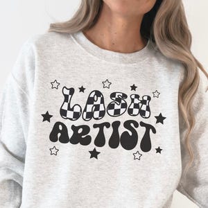 Peut inclure: Un sweat-shirt gris clair à col rond avec le texte noir "LASH ARTIST" écrit dans une police ondulée d'inspiration rétro. Le mot "LASH" présente un motif à damier et le design est entouré de plusieurs petites étoiles noires de tailles variées.