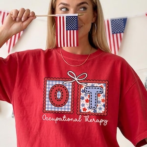 May include: A red t-shirt with the letters "OT" in a postage stamp design, with the words "Occupational Therapy" underneath. The shirt is worn by a person holding a small American flag. Patriotic decorations are in the background.
