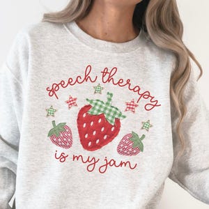 May include: An ash gray crewneck sweatshirt featuring a red strawberry graphic with a green gingham leaf, smaller strawberries, and red stars. The text reads "speech therapy is my jam" in red cursive script. The fabric has a heathered texture.