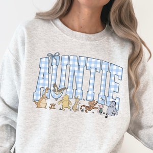 May include: A light gray sweatshirt with the word "AUNTIE" in large, blue gingham letters. Below the text are cartoon characters. The sweatshirt is a comfortable, casual style.