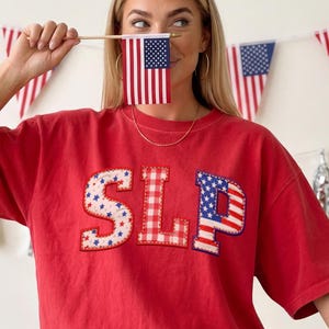 May include: Red t-shirt with the letters "SLP" appliqued on the front. The "S" is white with blue stars, the "L" is red and white checkered, and the "P" is a US flag design. The person is holding a small American flag.