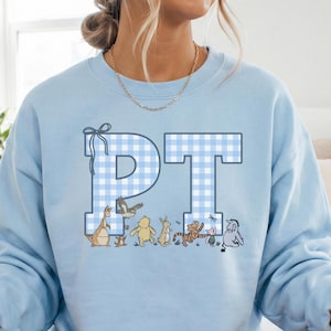 May include: A light blue crewneck sweatshirt featuring the letters PT in a white and blue gingham pattern. Below the letters are small illustrations of various cartoon characters including a bear, a tiger, a piglet, and a donkey.