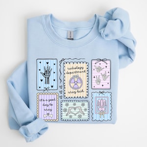 May include: Light blue sweatshirt with multiple framed illustrations. The illustrations include a hand, the words "radiology department" and "xray tech", a heart made of flowers, and a skeleton. The sweatshirt has the text "It's a good day to xray".
