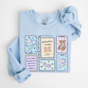 May include: Light blue sweatshirt with a colorful graphic design. The design includes illustrations of a stethoscope, teddy bear, band-aid, and the words "pediatric nurse" and "it's a good day to care for tiny humans."