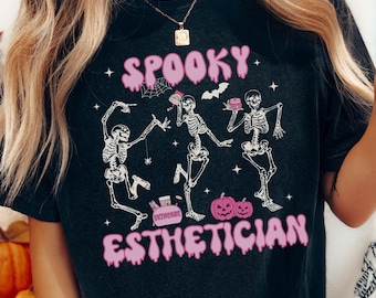 May include: A black short-sleeve t-shirt featuring a graphic print of three white skeletons dancing. The text reads SPOOKY ESTHETICIAN in pink bubbly letters. The design includes small pink pumpkins, a skincare bag, and tiny stars.