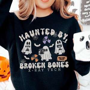 May include: A black long-sleeve sweatshirt featuring a graphic print with three white ghosts, spiderwebs, bats, and radiation symbols. The text reads "HAUNTED BY BROKEN BONES X-RAY TECH" and "HOLD YOUR BREATH" in a cream-colored, retro-style font.