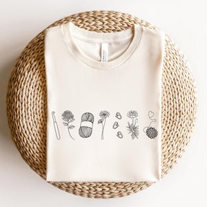 May include: A white t-shirt with a black line drawing of a crochet hook, yarn, and flowers.