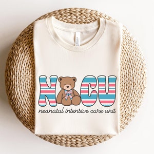 Puede incluir: Una camiseta de color crema con un gr&aacute;fico de un osito de peluche en el centro de las letras "NICU" en rayas azules, rosas y blancas. El texto "unidad de cuidados intensivos neonatales" est&aacute; debajo del gr&aacute;fico.