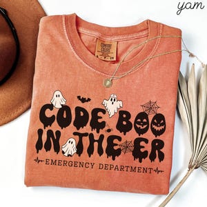 May include: A terracotta-colored t-shirt featuring a black graphic with the text CODE BOO IN THE ER and EMERGENCY DEPARTMENT. The design includes illustrations of ghosts, a bat, spiderwebs, and a jack-o'-lantern, with a heartbeat line graphic.