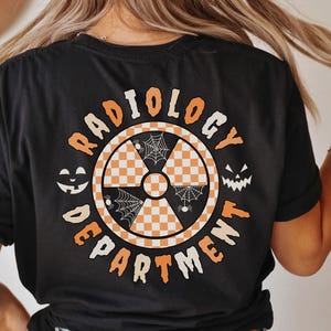 May include: A black t-shirt featuring a circular graphic on the back with the text RADIOLOGY DEPARTMENT in orange. The center contains a radiation symbol with a white and orange checkerboard pattern, spider webs, and two jack-o'-lantern faces.