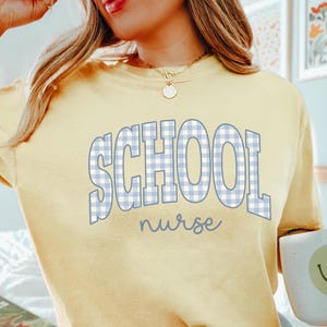 May include: Yellow sweatshirt with the words "SCHOOL nurse" in blue gingham print. The sweatshirt is a light yellow color and has a relaxed fit. The text is in a playful font, making it suitable for school nurses.