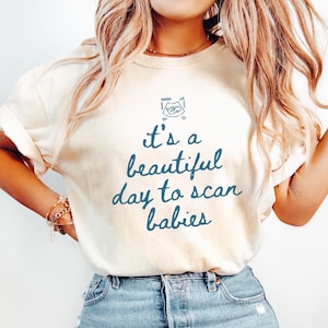 May include: Ivory-colored t-shirt with the text "it's a beautiful day to scan babies" in blue script. The shirt features a small graphic above the text. The model is wearing blue jeans.