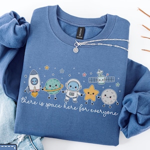 May include: An indigo crewneck sweatshirt featuring a graphic of a rocket, a planet with rings, an astronaut, a star, a satellite, and a moon, all with smiling faces. The text "there is space here for everyone" is printed in white cursive script.