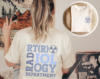 May include: A cream colored t-shirt with the text "RT(R) RADIOLOGY DEPARTMENT" in blue and a blue radiation symbol. The text is stacked vertically with the words "RAD" and "OLOGY" in a larger font.