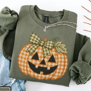 May include: A military green sweatshirt featuring a large applique of a jack-o'-lantern pumpkin with an orange and white gingham pattern. The pumpkin has black felt eyes and a mouth, topped with a green and white gingham bow and a fabric leaf.
