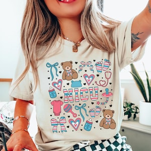 May include: A light beige t-shirt with a graphic design of a pink and blue baby footprint, a stethoscope, a teddy bear, a rattle, a pacifier, a bottle, a bow, and the word "NICU" in pink and blue letters.