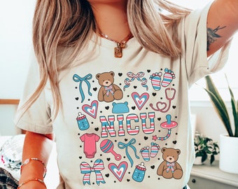 May include: A light beige t-shirt with a graphic design of a pink and blue baby footprint, a stethoscope, a teddy bear, a rattle, a pacifier, a bottle, a bow, and the word "NICU" in pink and blue letters.