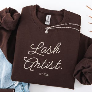 Peut inclure: Un sweat-shirt à col rond marron chocolat foncé avec le texte Lash Artist en police cursive beige clair, et le texte EST. 2026 imprimé en dessous dans une police sans empattement plus petite. Une chaîne en argent repose sur l'encolure.