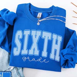 May include: A royal blue crewneck sweatshirt featuring the word SIXTH in large, light blue and white striped block letters with a scalloped outline, and the word grade written below in a white cursive script font, paired with blue denim jeans.