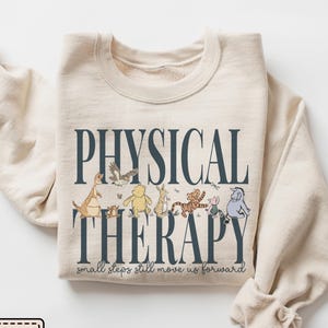 May include: A sand-colored crewneck sweatshirt featuring dark blue text that reads PHYSICAL THERAPY and small steps still move us forward. The design includes illustrations of various cartoon animal characters walking in a line across the center.