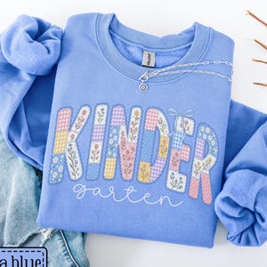 May include: A Carolina blue crewneck sweatshirt featuring the word KINDER in large, blocky letters filled with a patchwork pattern of floral and gingham prints, with the word garten written below in white cursive script and a small bow.