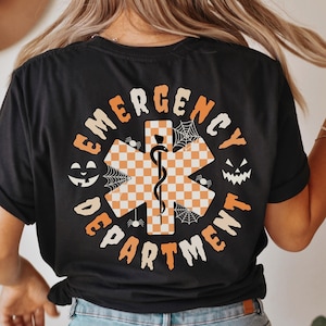 May include: Black t-shirt with an orange and white checkered star of life design, the words "EMERGENCY DEPARTMENT" in a circular pattern, and Halloween-themed accents like jack-o'-lanterns and spiderwebs.