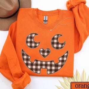 May include: A bright orange long-sleeve sweatshirt featuring a jack-o'-lantern face design. The eyes, heart-shaped nose, and jagged mouth are appliqued with a black and white buffalo plaid fabric. A brown hat is positioned near the top left corner.