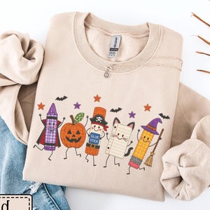 May include: A sand-colored crewneck sweatshirt featuring a graphic print of five anthropomorphic school supplies dressed in Halloween costumes, including a crayon, a pumpkin, a glue stick, a notebook, and a pencil, surrounded by small bats and stars.
