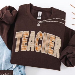 May include: A dark chocolate-colored sweatshirt with the word "TEACHER" in a patchwork design. The letters are a mix of plaid and floral patterns in orange and beige. A silver necklace with a small pendant rests on the sweatshirt. The sweatshirt is paired with blue jeans.